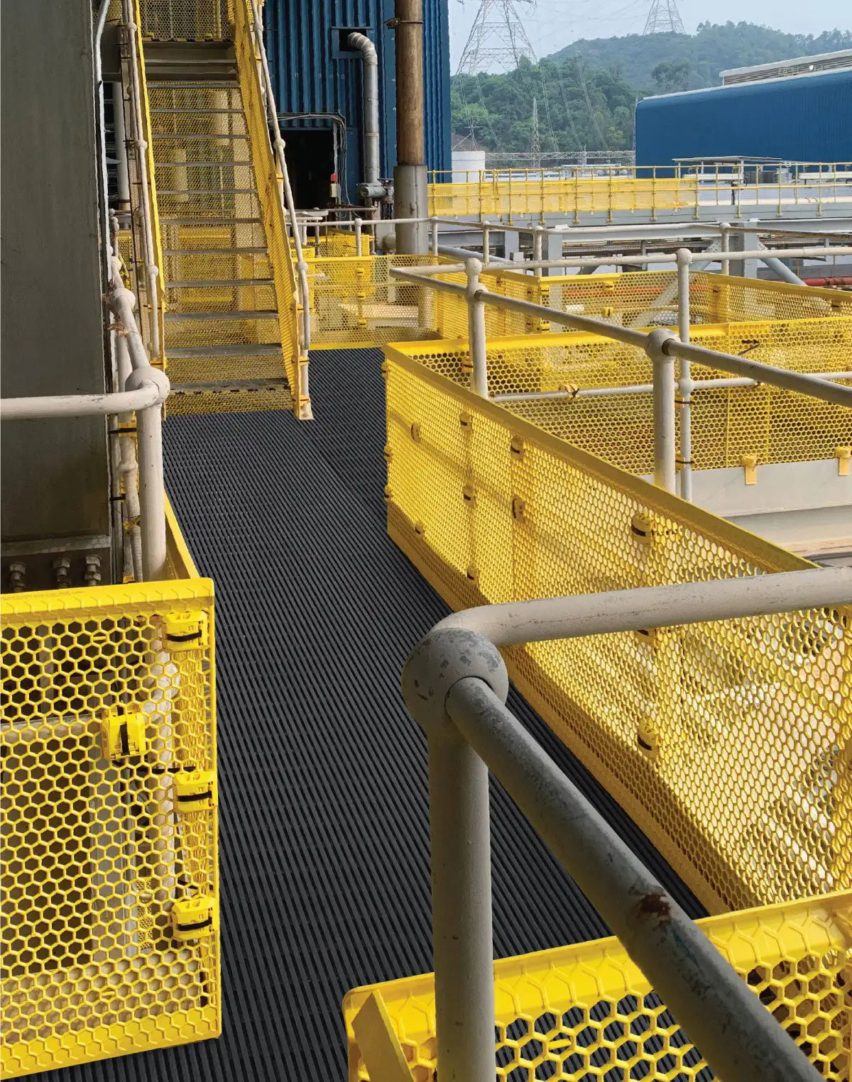 Black textured safety mat with yellow hexagonal mesh railings and industrial piping in the background.