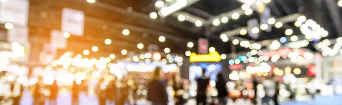 Blurry indoor exhibition hall with bright overhead lights and indistinct figures in the crowd.