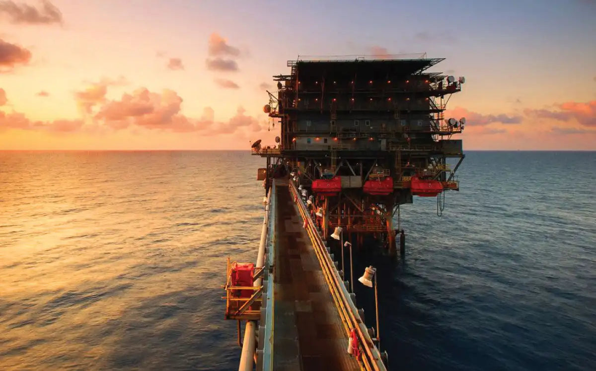 A massive offshore oil rig with a dark metallic structure, red safety equipment, and illuminated lights against a sunset sky.