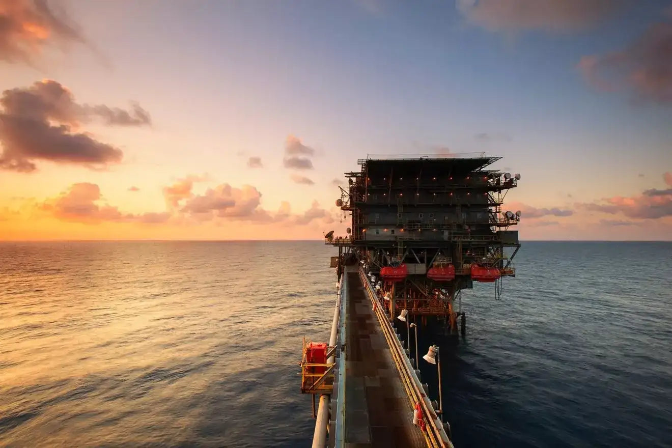 A massive offshore oil rig with a dark metallic structure, red safety equipment and walkways extending into the ocean under a sunset sky.