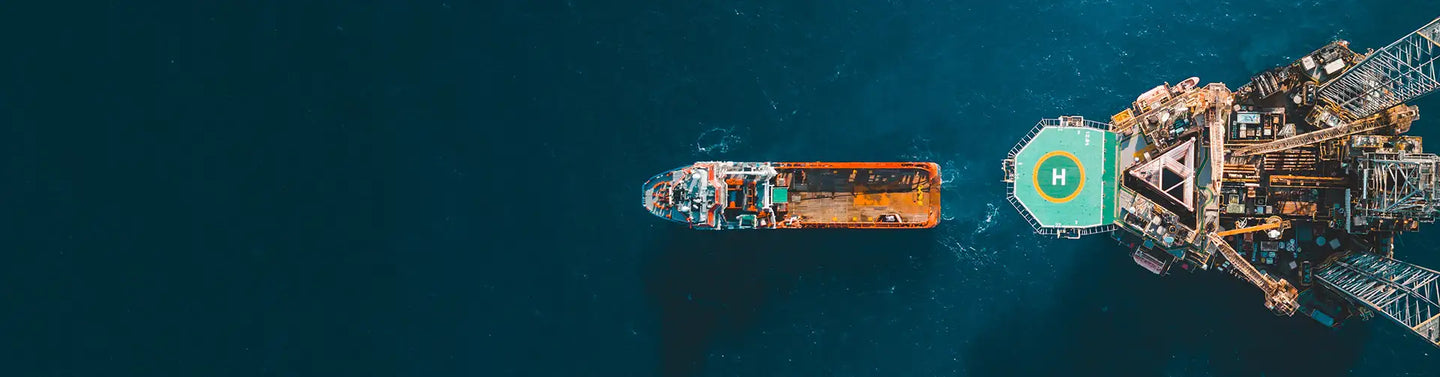 A rusted orange and white oil rig platform with a green helipad marked by a central ’h’ sits beside an industrial vessel in deep blue water.