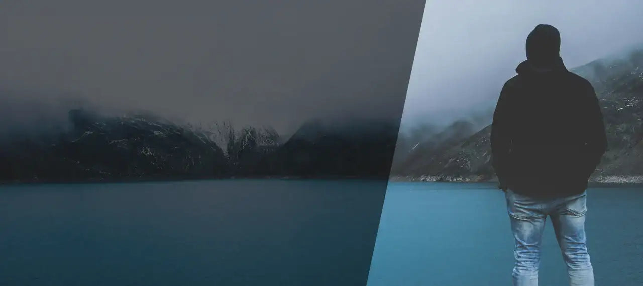 A solitary figure in a dark hooded jacket and light-wash jeans stands facing away, gazing out over a misty lake with distant mountains shrouded in fog.