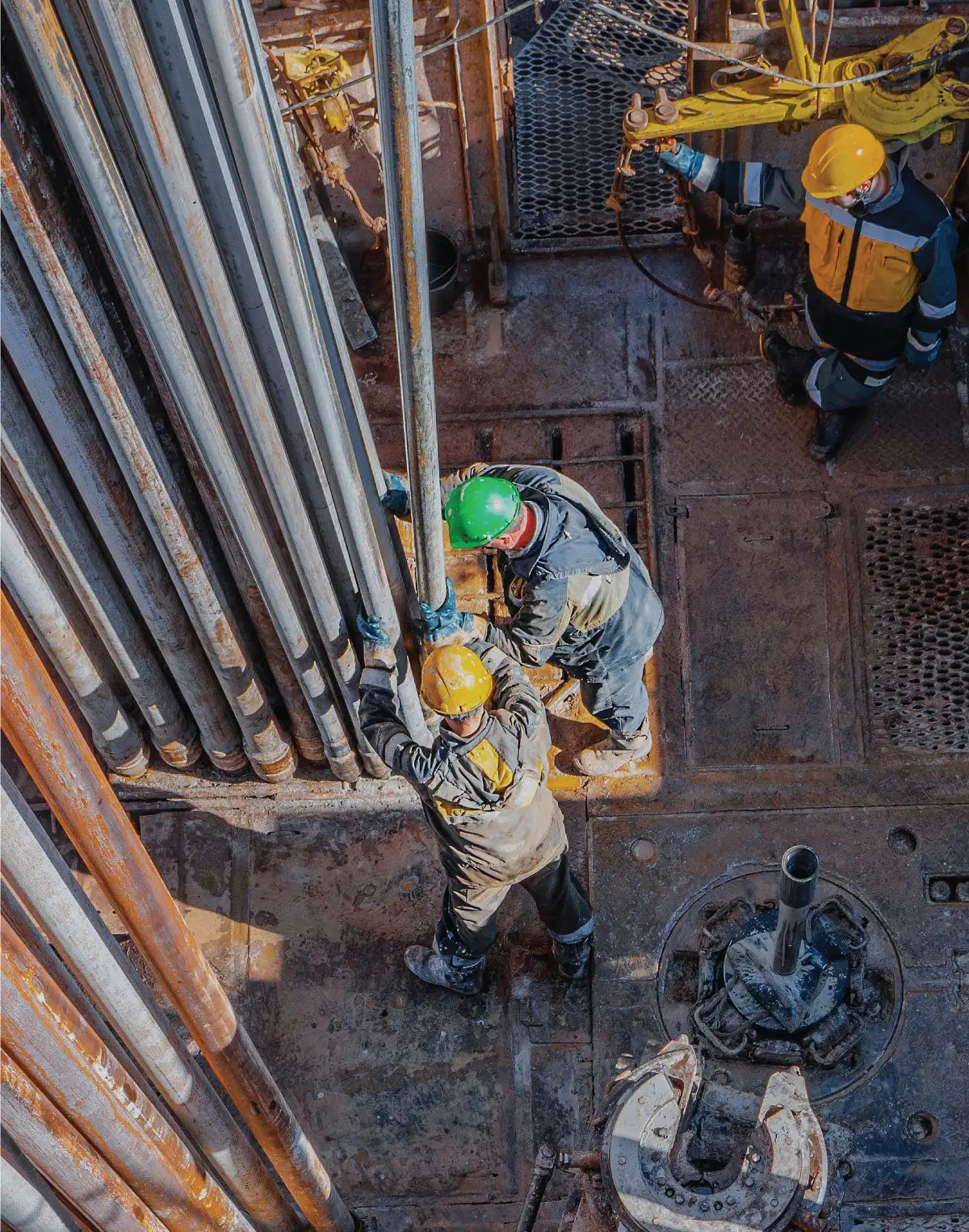 Three workers in hard hats and work gear are handling thick, metallic drill pipes on an industrial rig.