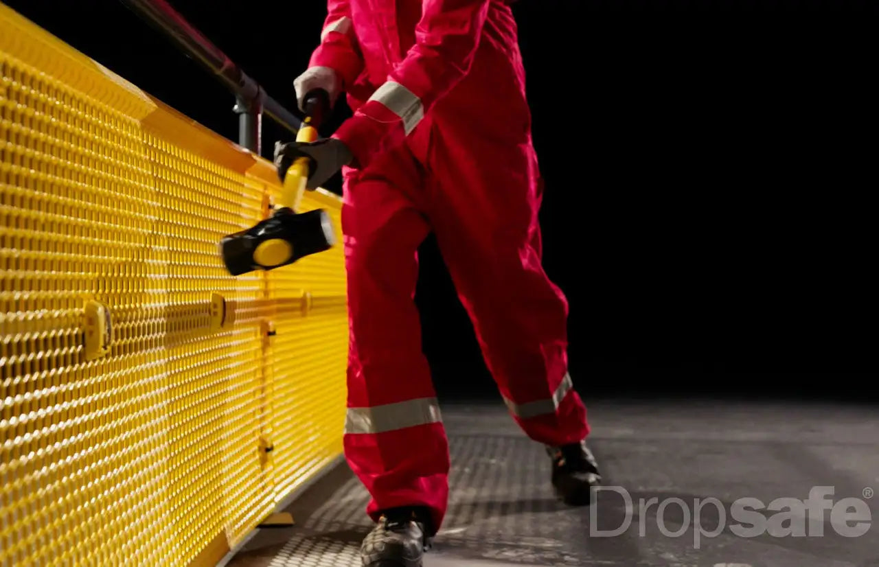 A worker in a red safety jumpsuit holds a yellow and black sledgehammer, poised to strike the bright yellow perforated metal barrier.